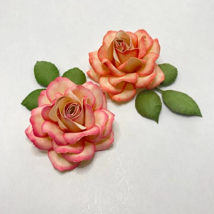 Duas rosas de papel em tons de pêssego com borda rosa e folhas verdes